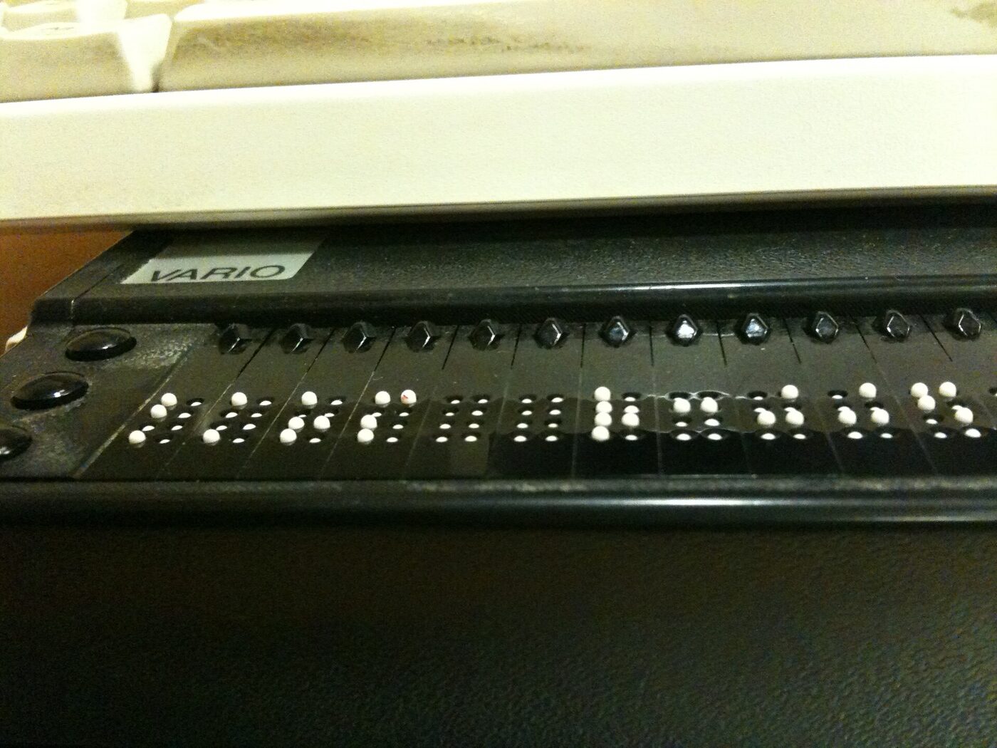 Close-up of a black braille display beneath the front edge of a keyboard, its row of cells showing raised white dots, with small routing buttons above each cell.