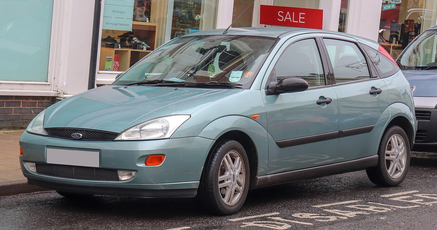 Front three-quarter view of a pale green 1990s Ford Focus five-door hatchback with rounded body sides, tall triangular headlamps and an oval Ford badge on the grille, parked at a kerb outside a row of shops.