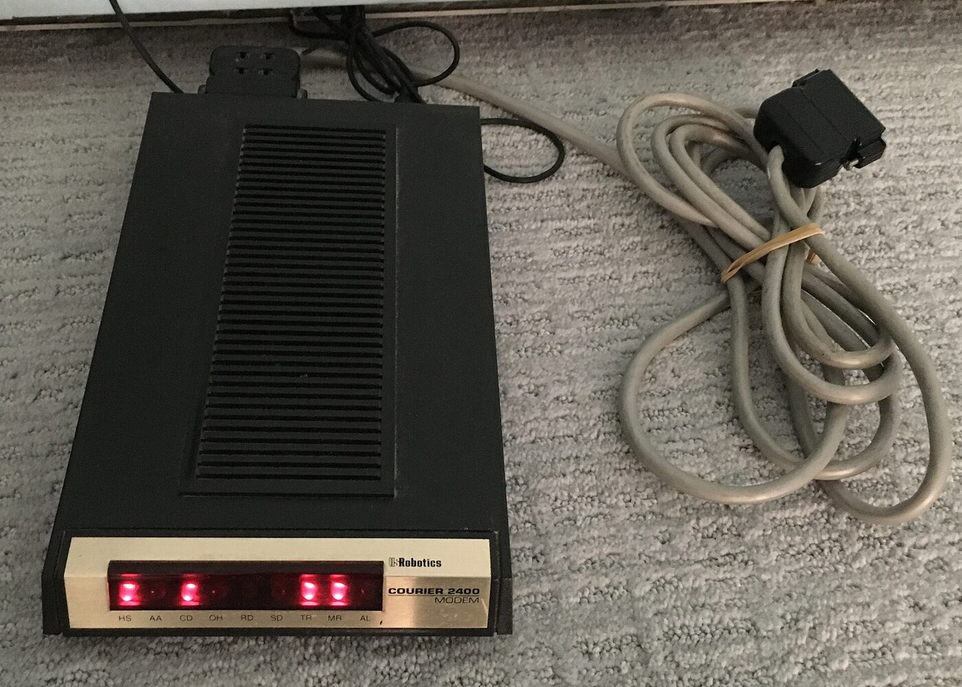 A black wedge-shaped desktop modem photographed from above on carpet, its front panel lettered USRobotics Courier 2400 and carrying a row of red indicator lamps labelled HS, AA, CD, OH, RD, SD, TR, MR and AL, several of them lit. A coiled grey serial cable with a 25-pin connector lies beside it.
