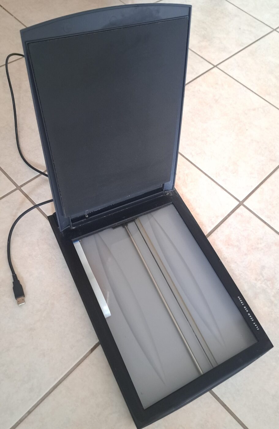 An open consumer flatbed scanner lying on a tiled floor, its hinged lid raised to show the glass platen and a trailing USB cable.