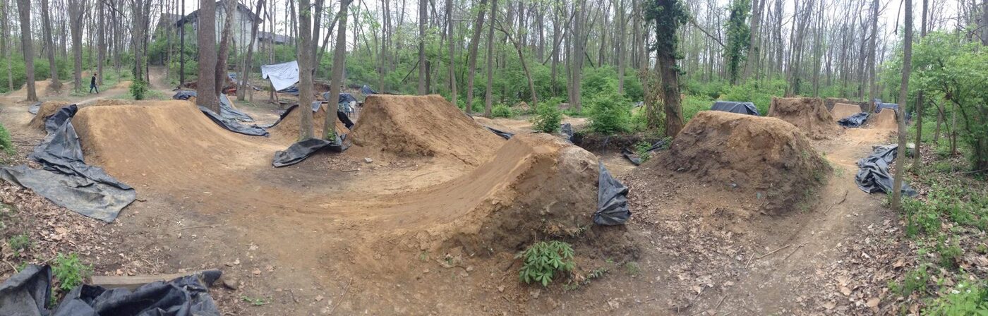 A panoramic view of a line of compacted-earth jumps in a wooded clearing, each with a steep take-off mound and a separate landing mound, with black plastic sheeting draped over some of the faces.