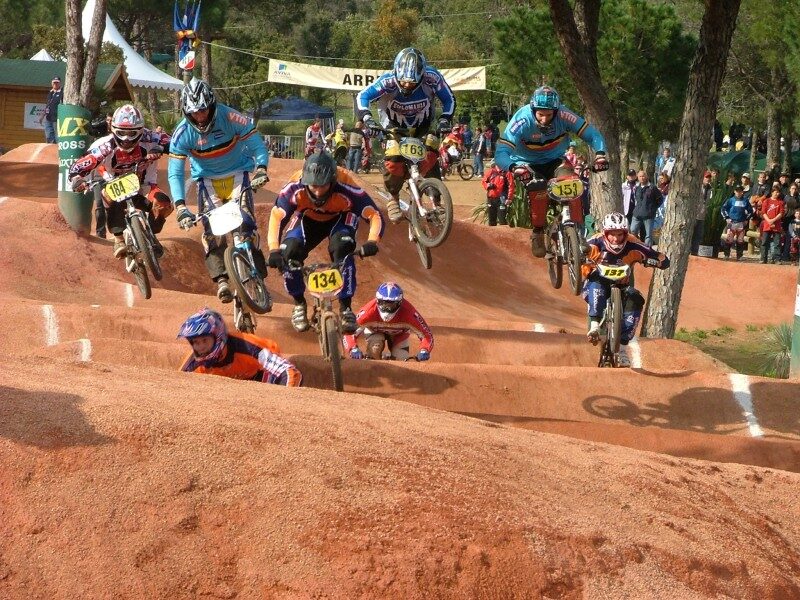 Eight BMX racers in helmets and full-face protection riding close together over a series of red-earth rollers at a race meeting, spectators lining the track behind them.