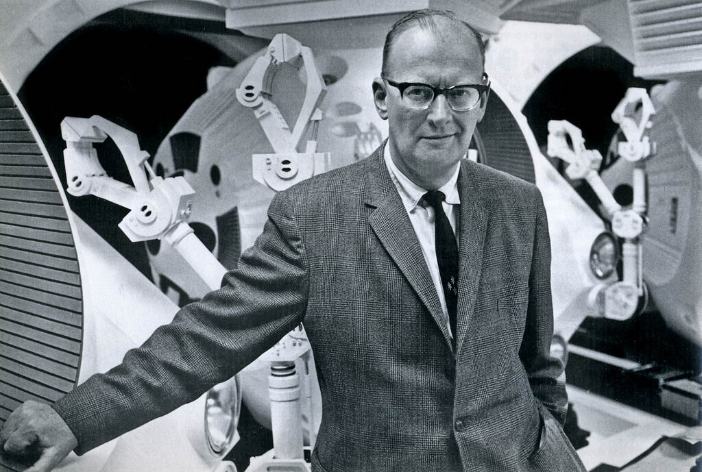 Black-and-white photograph of a man in middle age wearing heavy dark-rimmed glasses and a checked tweed jacket with a white shirt and dark tie, standing and looking towards the camera in front of large curved machinery fitted with jointed mechanical arms.