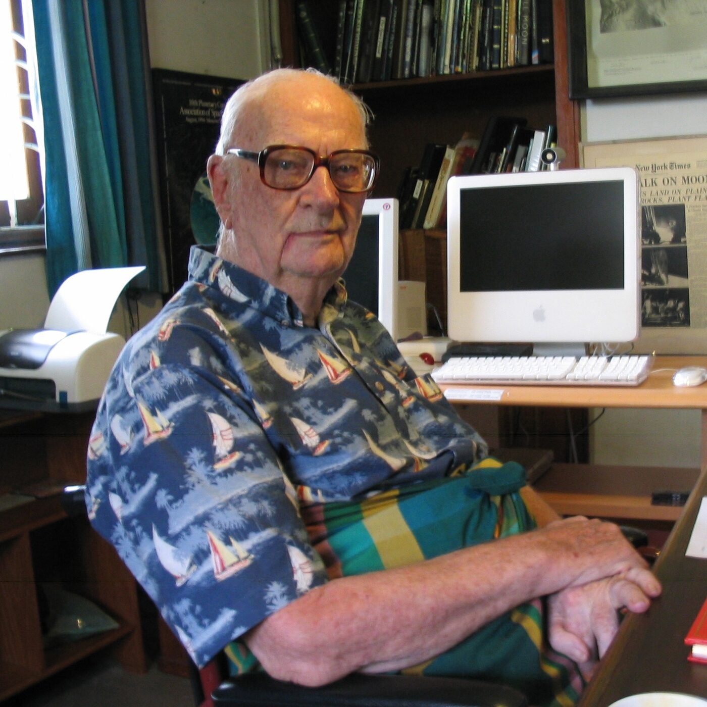 Colour photograph of an elderly man in a patterned short-sleeved shirt and a green and yellow sarong, seated at a desk in a book-lined study, with an Apple iMac, keyboard and mouse behind him and a mounted New York Times front page headlined about the Moon landing on the wall.