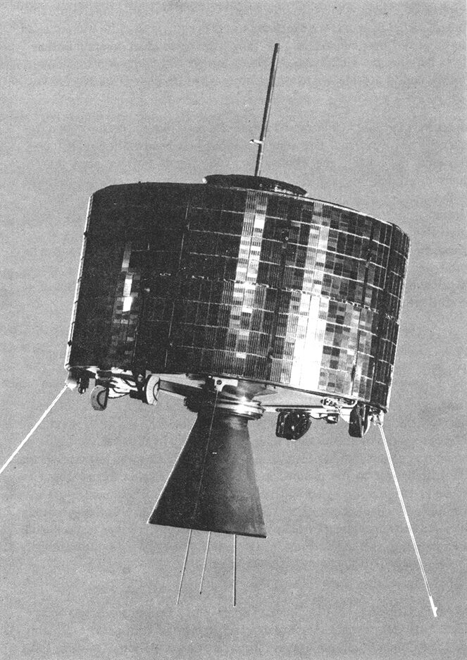 Black-and-white photograph of a squat drum-shaped satellite against a plain grey background, its cylindrical body covered in solar cells, with a slender antenna rod projecting from the top and a conical rocket nozzle and thin whip antennas below.