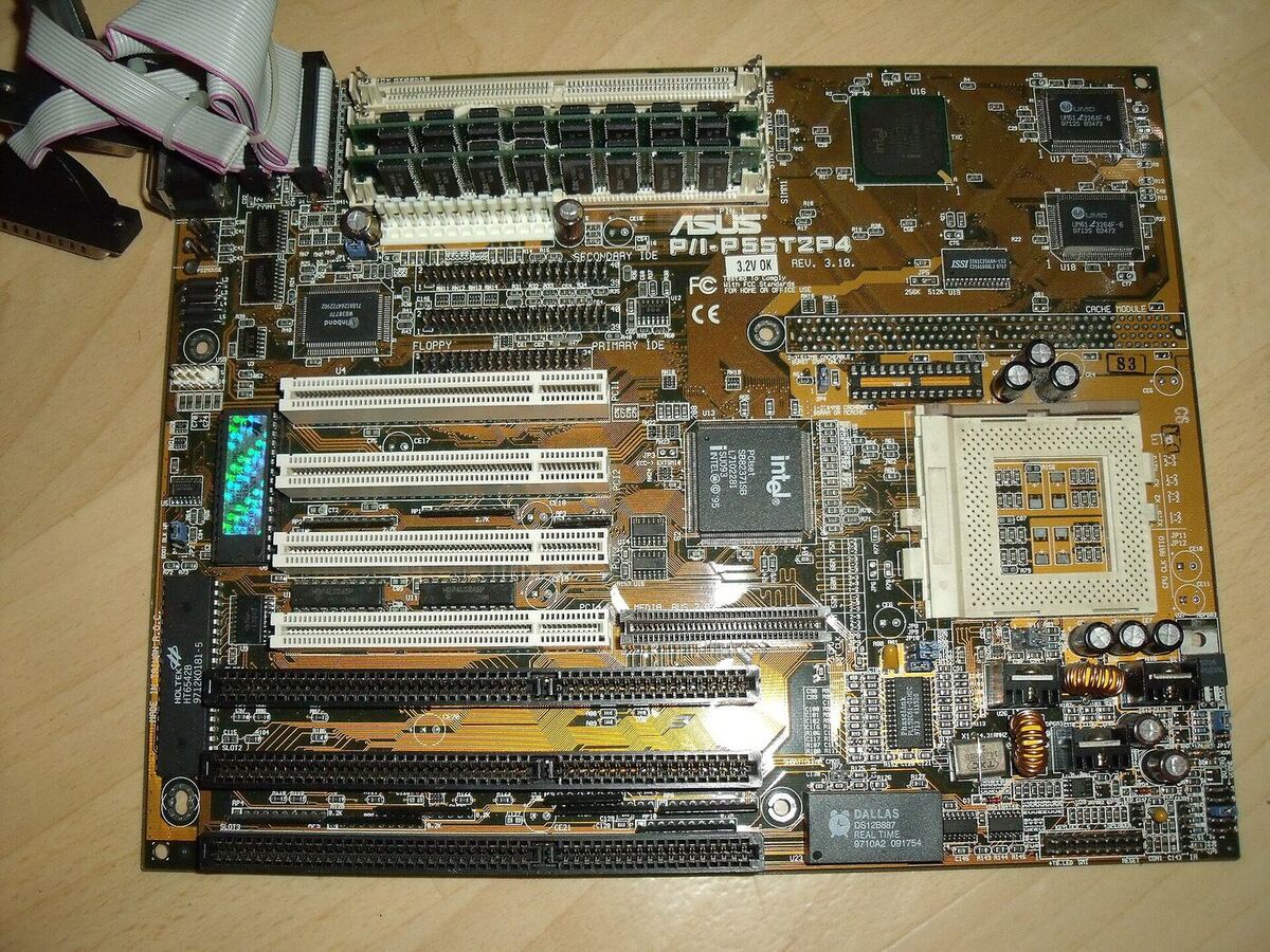 A brown printed circuit board photographed from above on a wooden table, with an empty white square processor socket at the right, four memory slots holding modules along the top, four white PCI slots and three longer black ISA slots, and an Intel chipset chip in the middle.