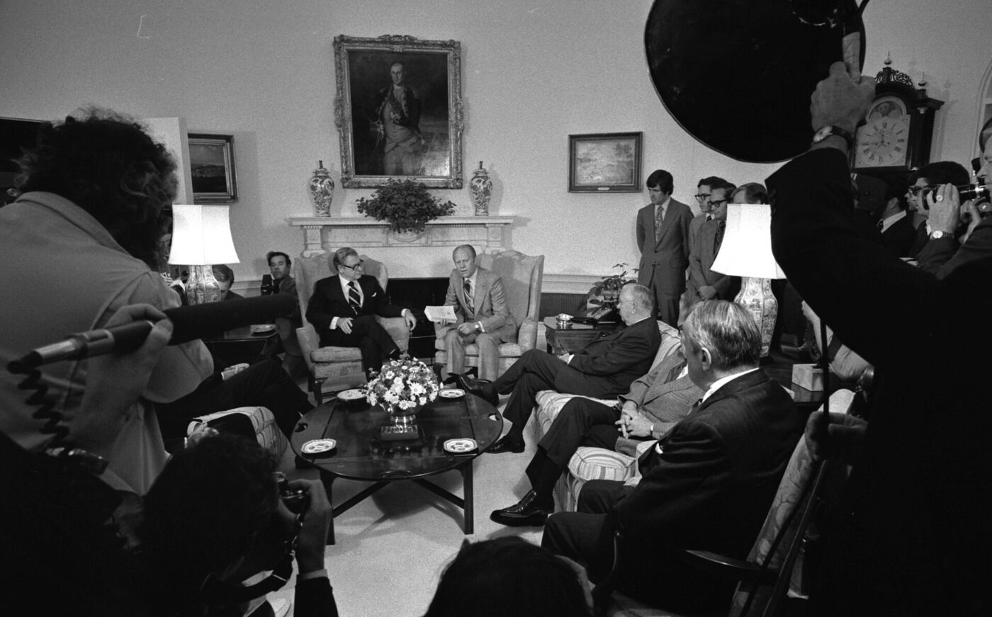 Black-and-white photograph of President Gerald Ford seated beside Vice-President Nelson Rockefeller in a White House sitting room, holding a printed report, surrounded by seated officials and a ring of press photographers.