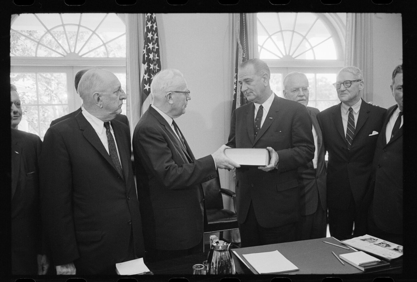 Black-and-white photograph of men in dark suits standing in a White House room, one of them handing a thick bound volume to President Lyndon B. Johnson.