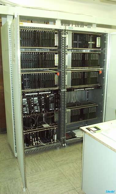 An open floor-standing equipment cabinet holding two bays of densely packed circuit cards, standing on a tiled floor beside a desk.