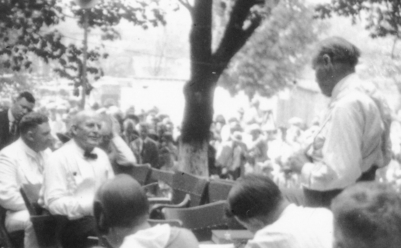 Black-and-white photograph of a court session held outdoors under a tree: an elderly man in shirtsleeves and bow tie sits at the left while a standing man in shirtsleeves faces him, with a large crowd of onlookers behind them.