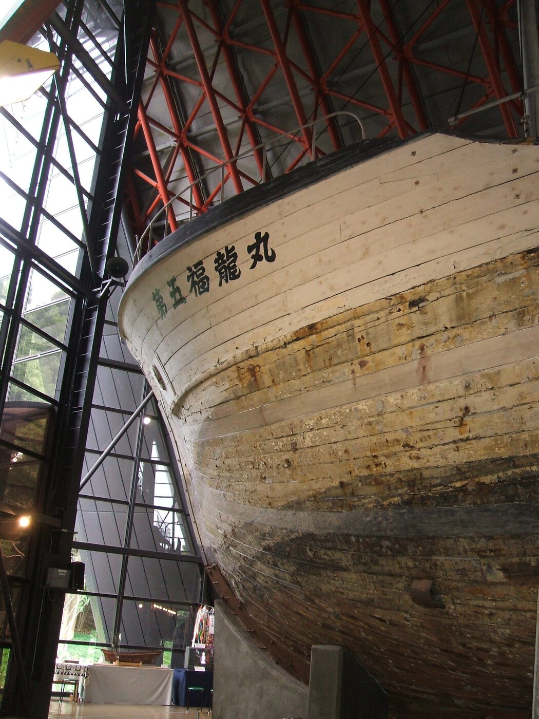 The bow of a wooden fishing boat preserved indoors, its Japanese name painted on the hull, under an exhibition hall roof.