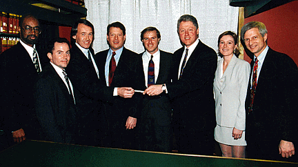 Group photograph of eight people in suits posed indoors; Bill Clinton and Al Gore stand among them as a pen is held out to a man in the centre.