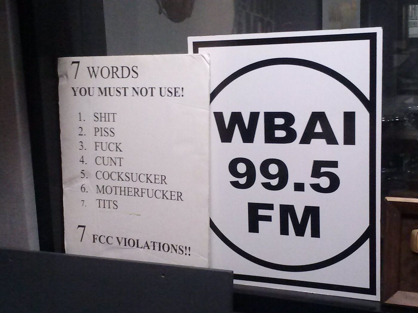 A printed notice in a radio studio headed "7 words you must not use", listing seven obscenities, beside a WBAI 99.5 FM logo sign.