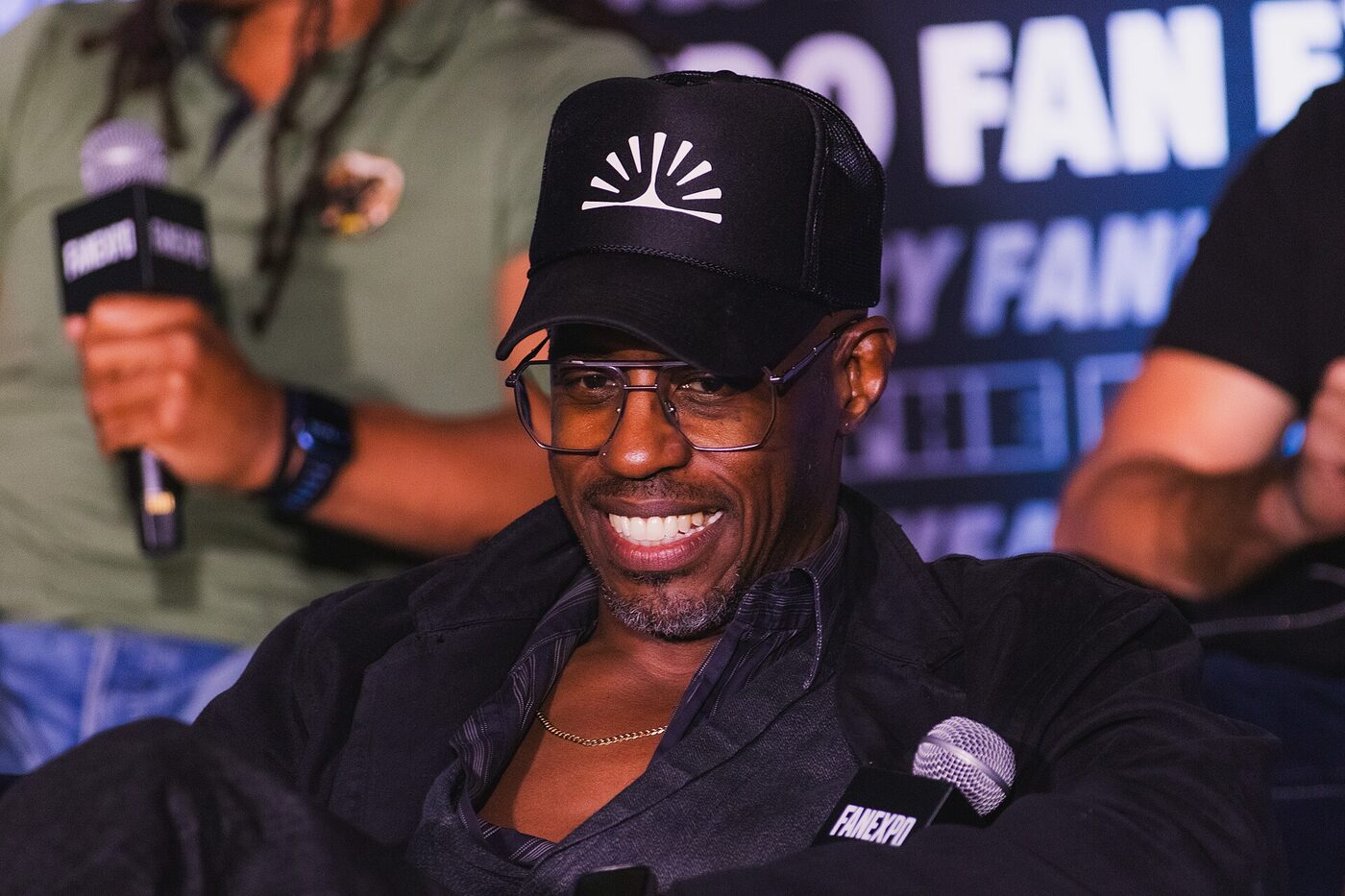 Ahmed Best, laughing, seated at a convention panel behind a microphone marked FANEXPO, wearing a black cap and wire-framed glasses, with another panellist and a step-and-repeat banner behind him.