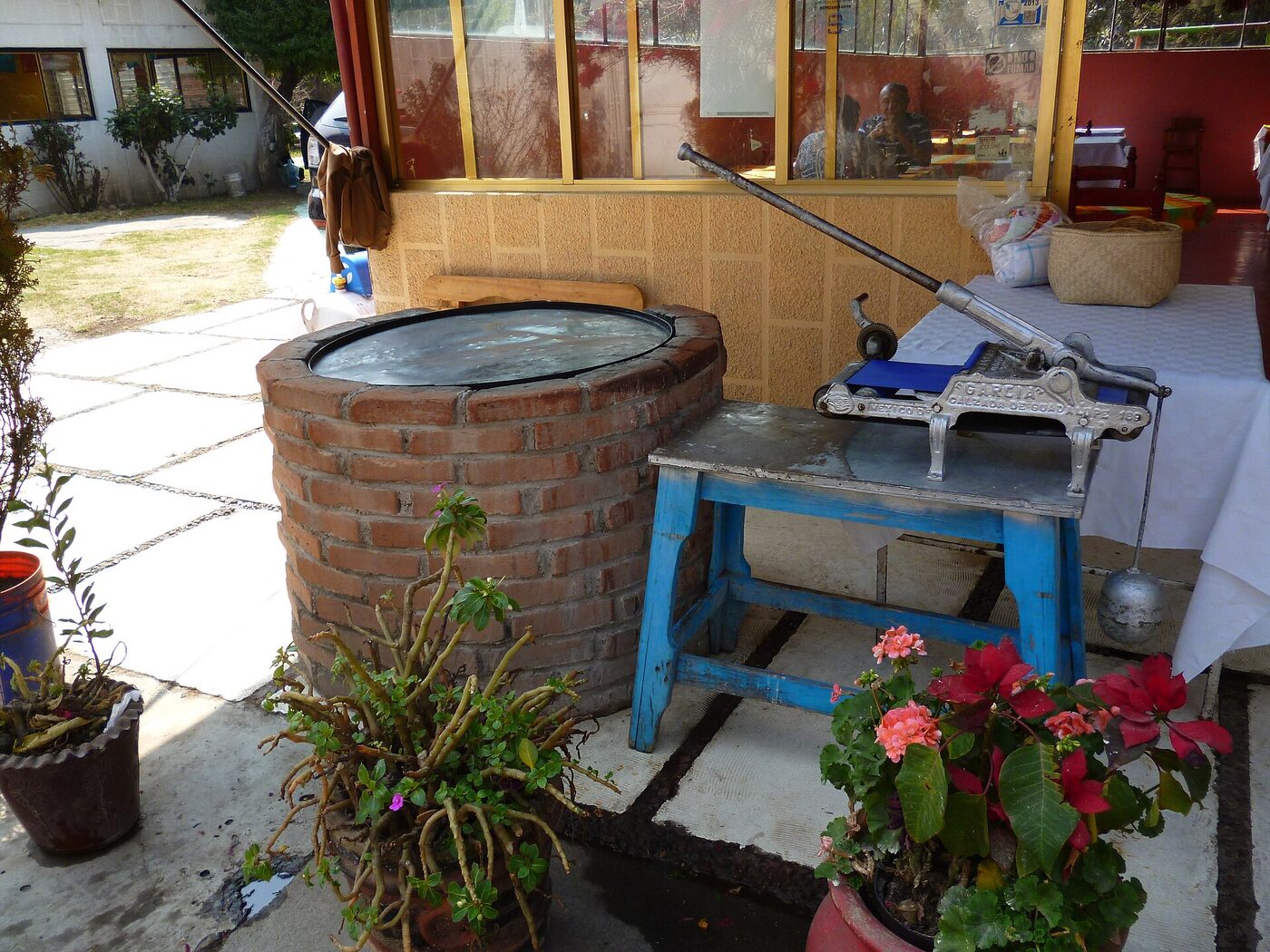 A large round metal griddle set into a circular brick base on an outdoor patio, with a heavy cast-iron lever tortilla press standing on a blue wooden table beside it.