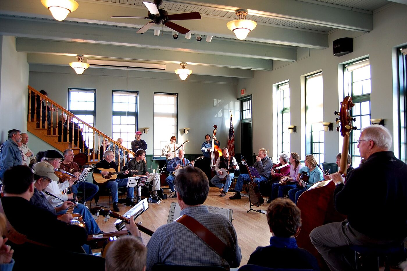 A group of seated musicians playing guitars, banjo, fiddle and upright bass in a circle in a plain indoor room.