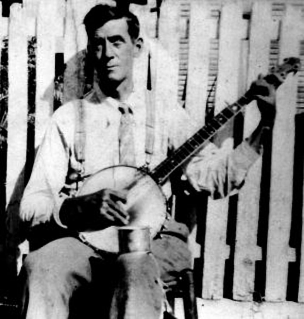 Sepia photograph of a man in a suit and hat standing before a picket fence, holding a banjo, with a cup hung round his neck.