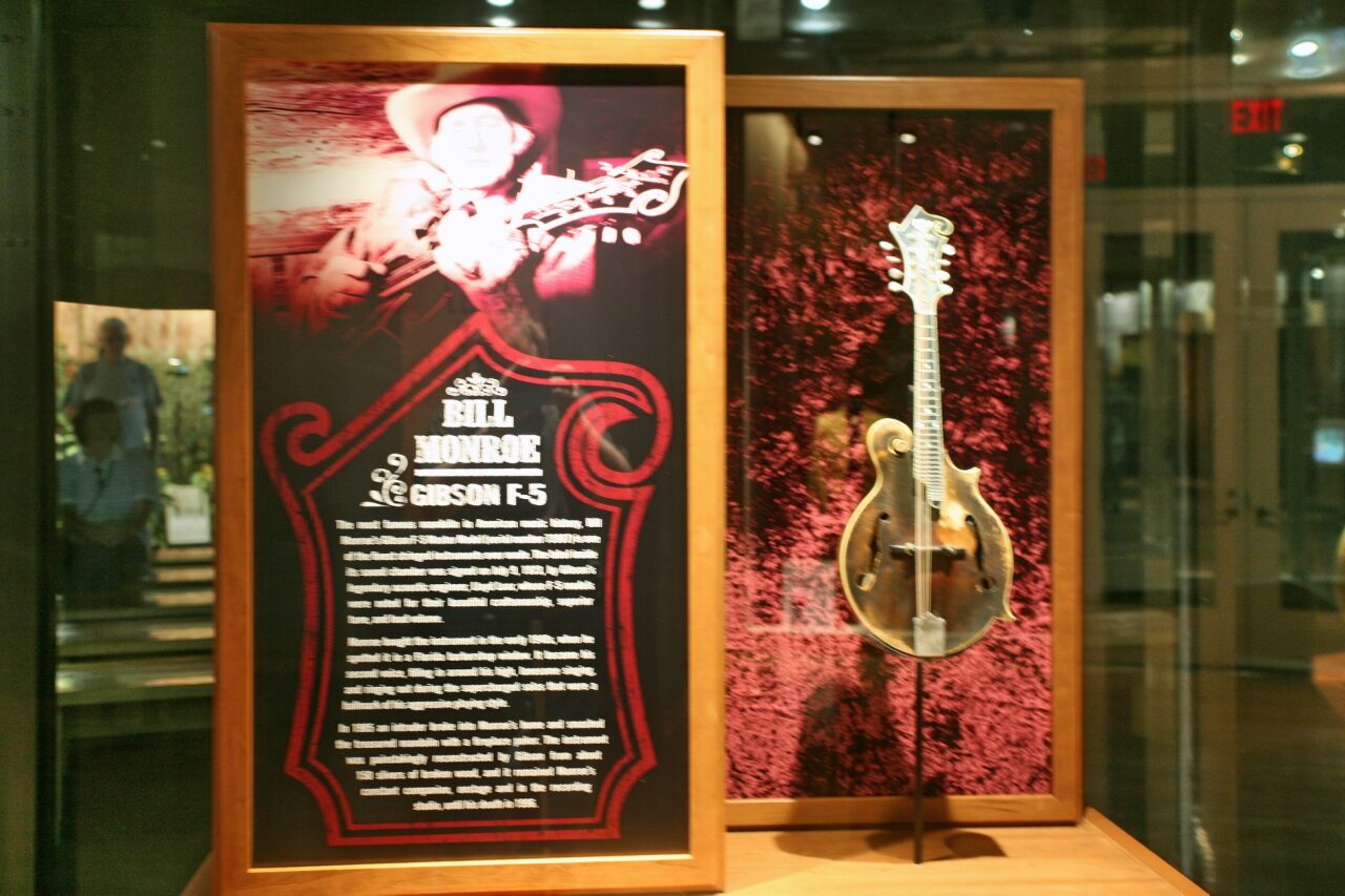 A carved-top mandolin with a scroll on the upper body, f-shaped sound holes and a sunburst finish, shown in a museum display.