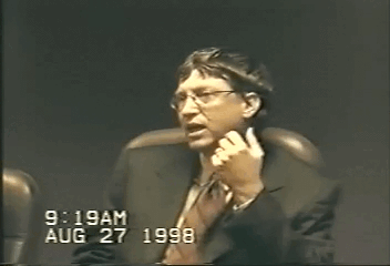 A grainy video still of a man in a dark suit and glasses seated in a chair, one hand raised as he speaks, with the timestamp 9:19AM AUG 27 1998 burned into the lower left corner.