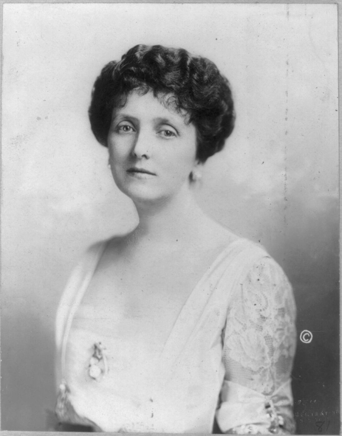 A monochrome 1912 studio portrait of Emily Post in a pale lace-sleeved dress, shown from the waist up.