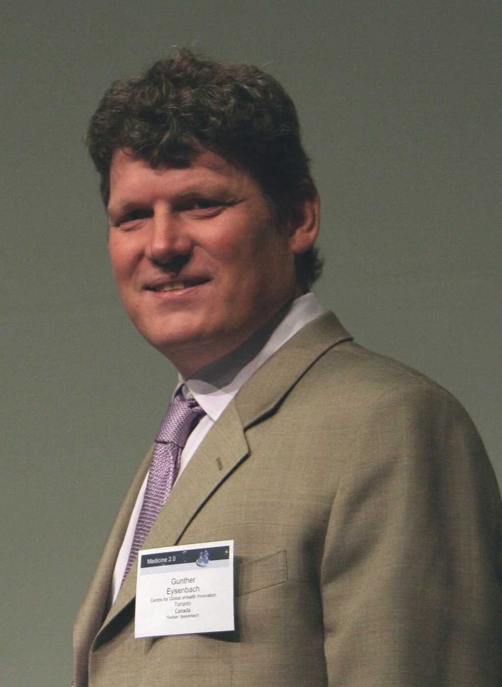 A man in a dark suit and open-collared shirt standing indoors at a conference, photographed from the chest up.