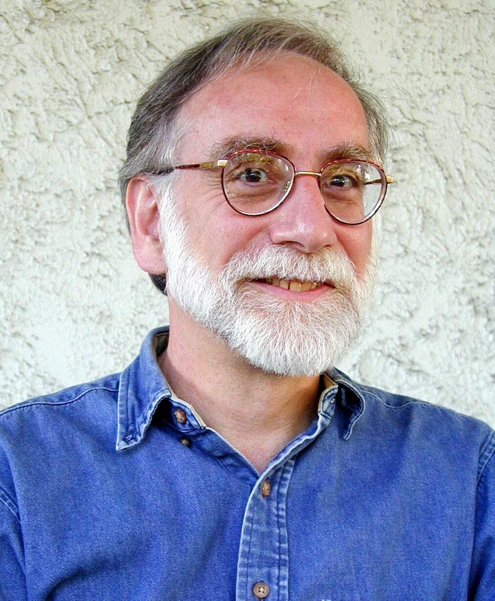 Head-and-shoulders portrait of a bearded man in glasses and an open-necked shirt, facing the camera indoors.