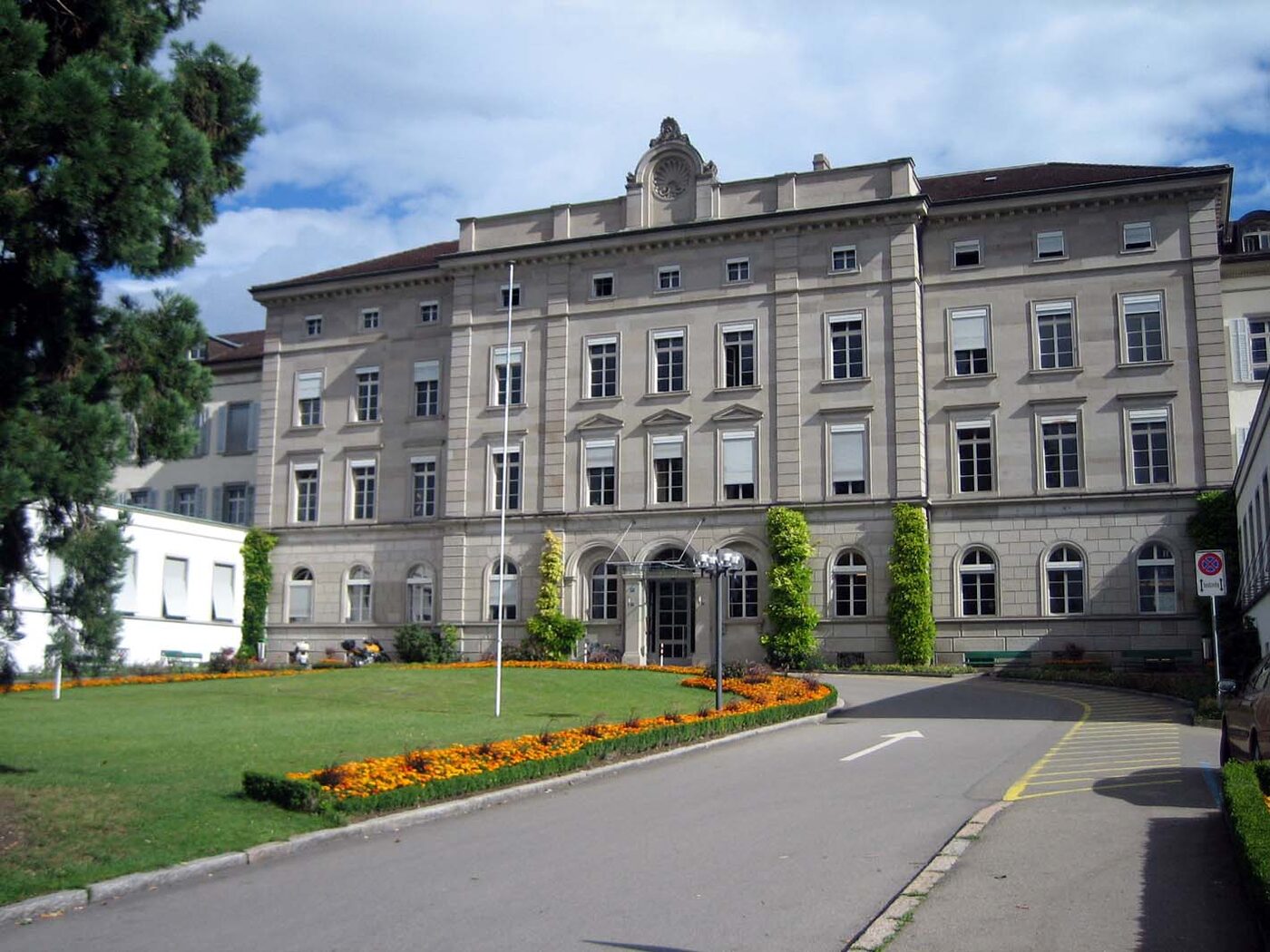 A large nineteenth-century institutional building of pale stone with a symmetrical facade, rows of tall windows and a central pedimented block, seen from the grounds on a bright day.
