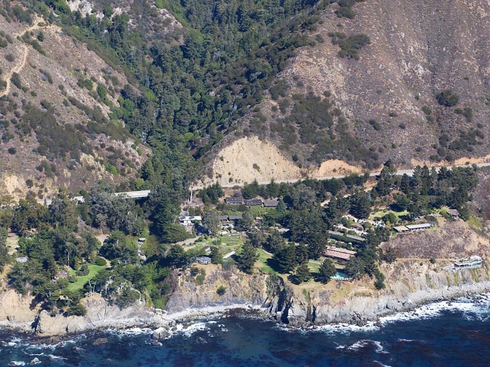 Aerial view of a scatter of low buildings, lawns and trees on a steep coastal terrace above the Pacific, with a highway cut into the hillside behind.