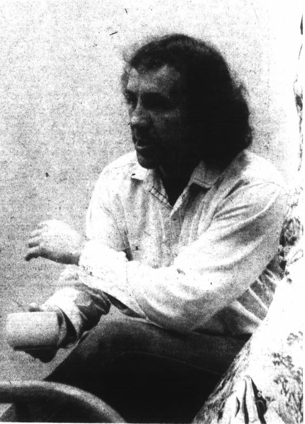 Grainy black-and-white photograph of a long-haired man in a light shirt, seated and talking, one hand raised mid-gesture.
