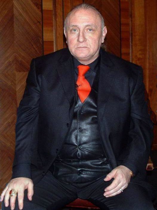 Colour photograph of a heavy-set older man with a receding hairline, seated in a black suit, black shirt and waistcoat with a bright red tie, against wood panelling.