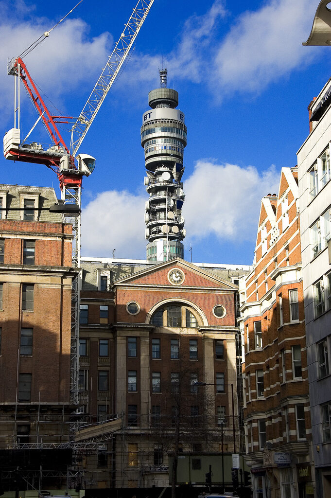 A brick hospital building with a pedimented clock frontage, seen down a London street between later buildings, with a construction crane at the left and the BT Tower rising behind it against a blue sky.