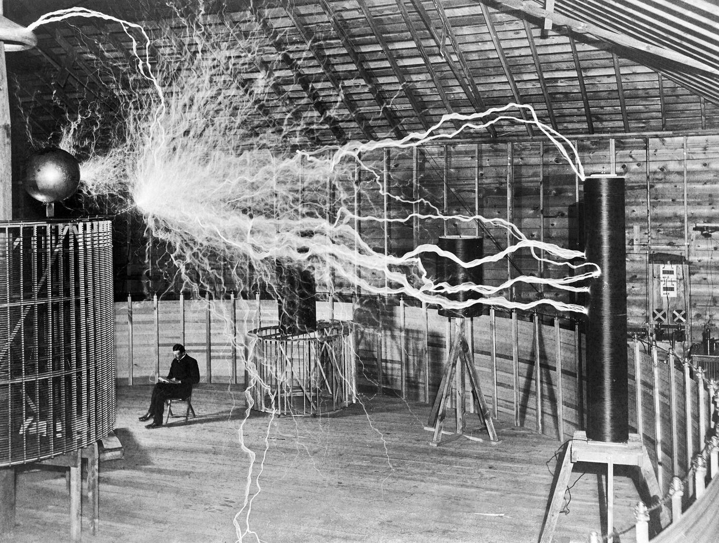 Black-and-white photograph of a large wooden laboratory interior in which enormous branching electrical arcs fill the upper space above a circular coil, while a man sits in a chair left of centre with an open book on his lap.