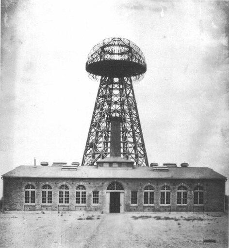 Black-and-white photograph of a tall open latticework tower carrying a broad ring-shaped lattice cupola at its top, standing behind a low single-storey brick building with arched windows, in flat open country.