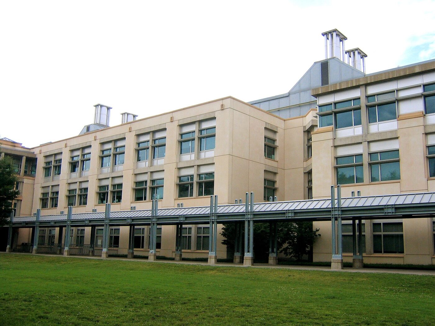 A long four-storey modern academic building in pale stucco and glass, with a covered walkway running along its ground floor and a lawn in front.