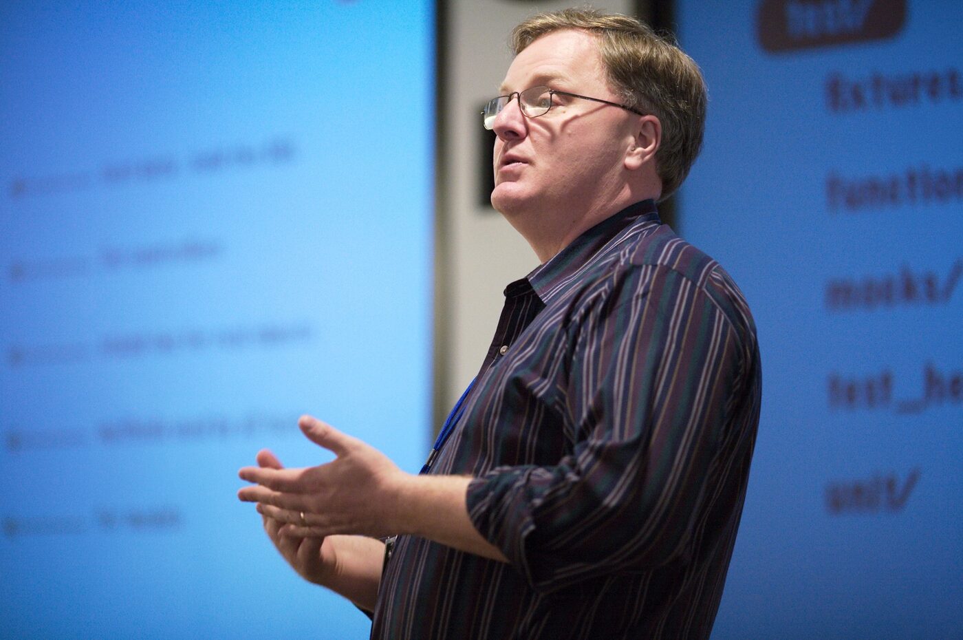 A bespectacled man in a striped shirt speaking with his hands raised, in front of a blue projection screen showing a bulleted slide.