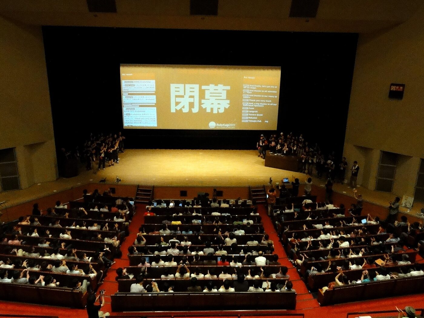 View from the back of a full auditorium, the audience applauding with raised hands, facing a stage and a large projection screen bearing two large Japanese characters.