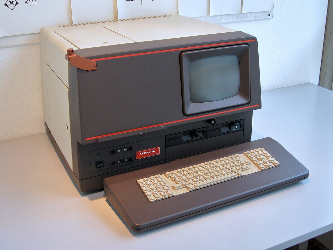 A desk-sized machine in grey and cream with a red trim line, a small monochrome screen at one end, two floppy-disk slots below it and a separate detached keyboard in front, standing on a white table.