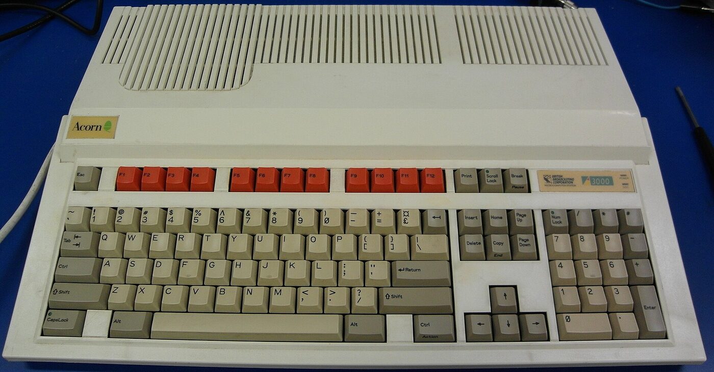 A beige wedge-shaped one-piece home computer with an integrated keyboard and red function keys, badged Acorn and A3000, resting on a blue bench.