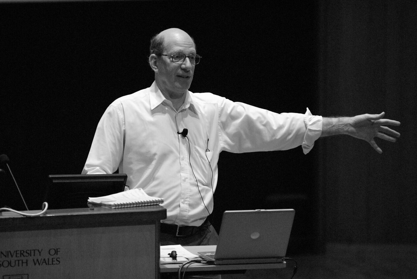Black-and-white photograph of a bespectacled man in a white shirt gesturing with one arm while speaking beside a lectern marked University of New South Wales, a laptop and papers on the desk in front of him.