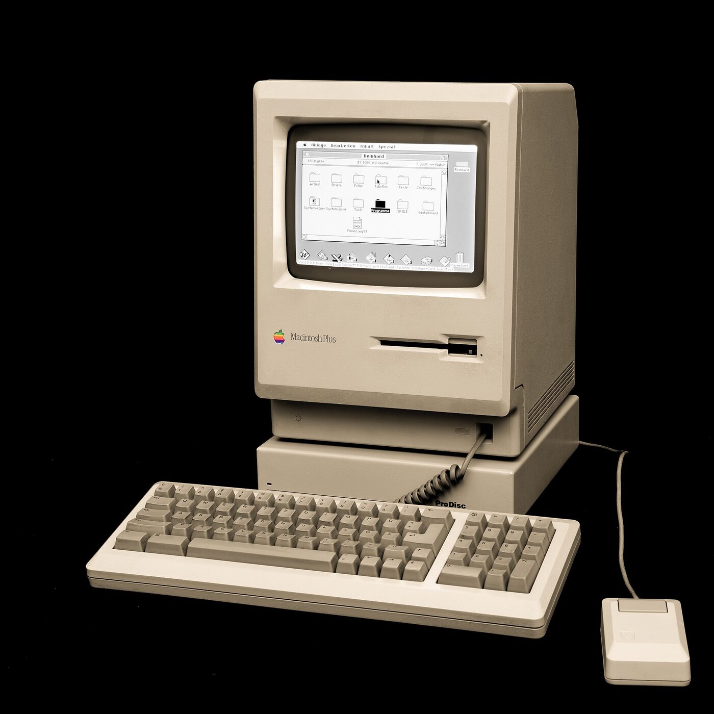 A beige all-in-one Macintosh Plus with a small built-in monochrome screen showing a file-manager window, sitting on an external disk unit, with a detached keyboard and a single-button mouse in front of it on a black background.