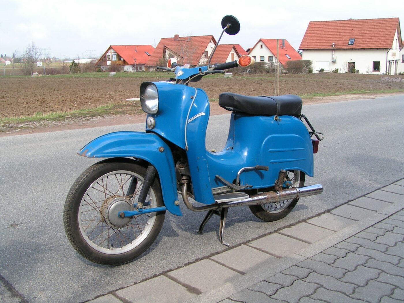 A blue East German motor scooter with a step-through frame, leg shield, single round headlamp and a low silencer on its left side, standing at a village roadside.