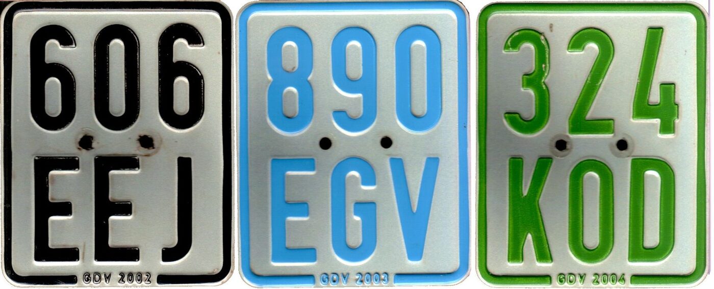 Three small upright vehicle plates on a pale reflective ground, each with three digits above three letters, lettered in black, blue and green respectively.