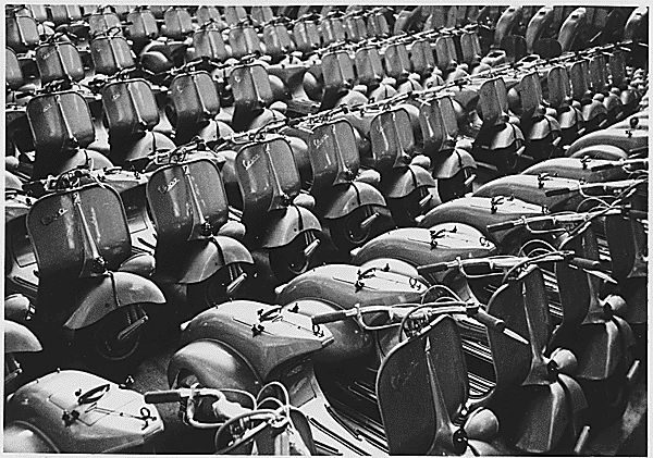 Black-and-white photograph looking down on dozens of newly built Vespa scooters parked in close rows at a factory.