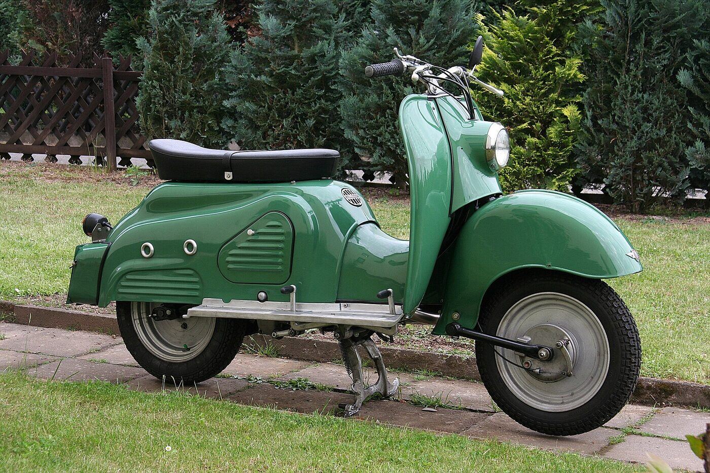 Side view of a green fully enclosed 1950s German motor scooter with large wheels, a deep front mudguard, a long bench seat and a flat floorboard.