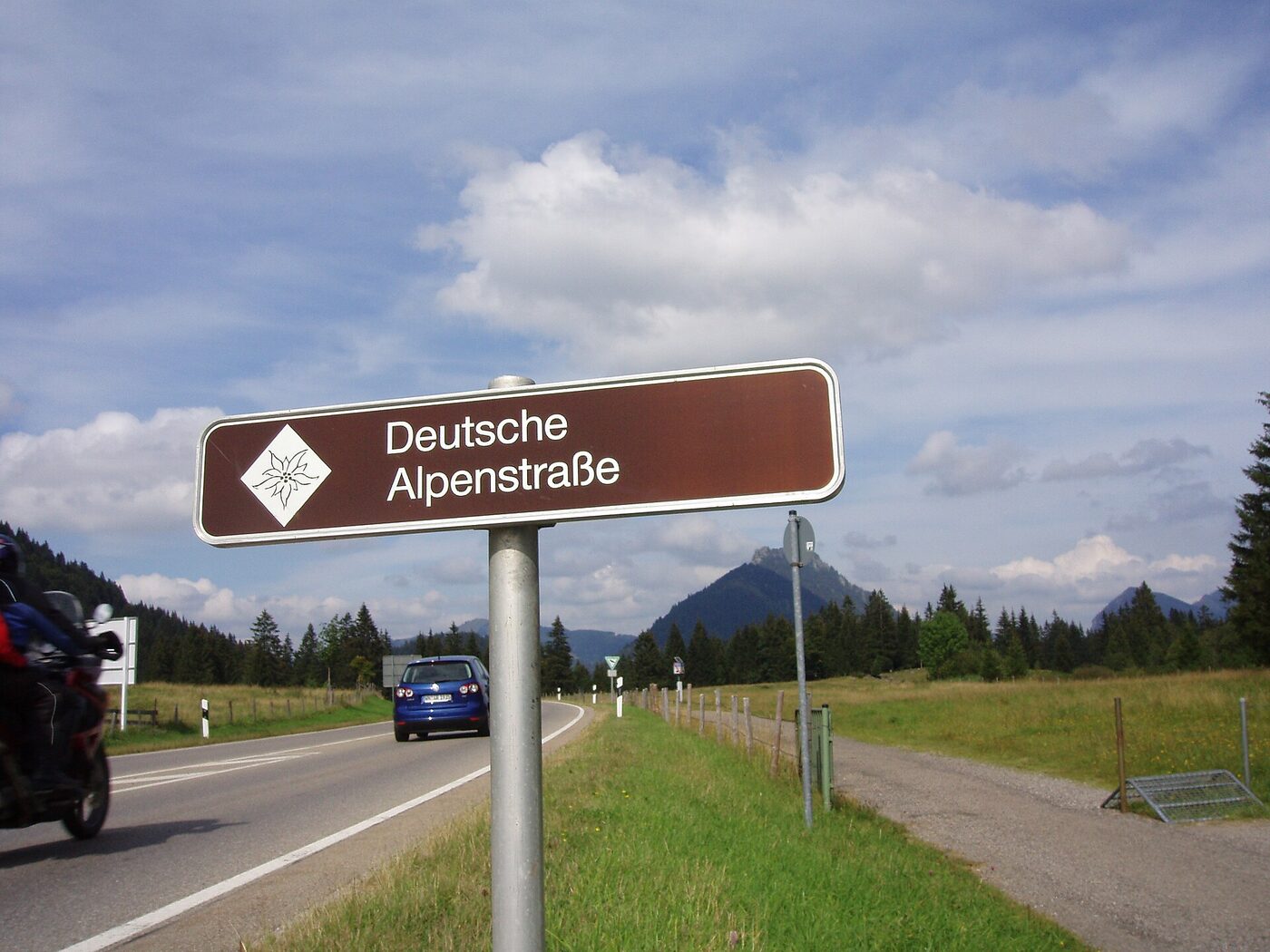 Brown roadside sign reading Deutsche Alpenstrasse with an edelweiss emblem, beside a two-lane road running through meadows towards wooded hills.