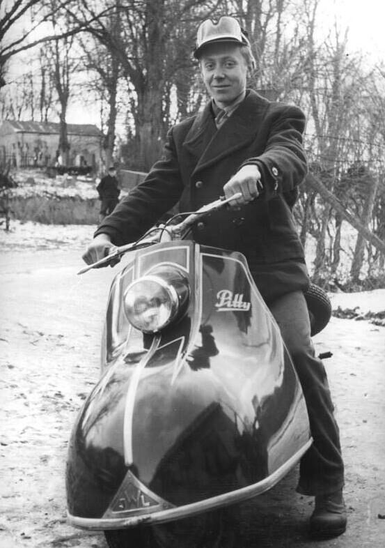 Black-and-white 1956 photograph of a young man in a coat and cap astride a Pitty motor scooter on a snow-covered country lane.