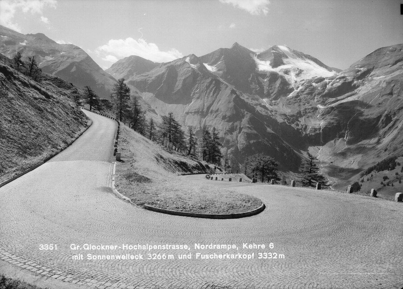 Black-and-white photograph of a cobbled hairpin bend on a mountain road, empty of traffic, with high rocky snow-streaked peaks behind.