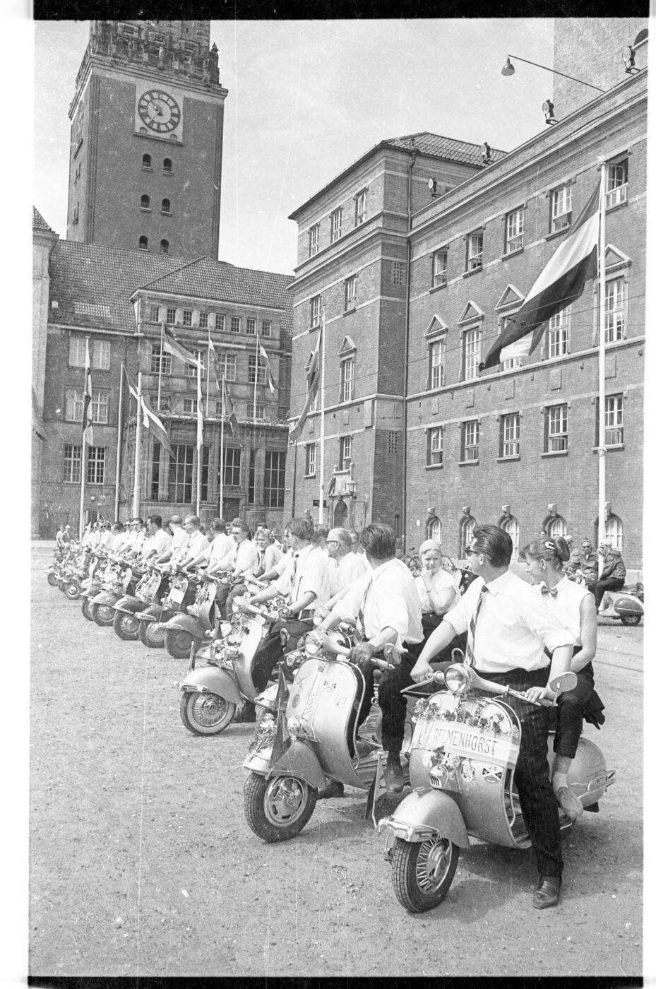 1957 photograph of a long line of Vespa scooters with riders and pillion passengers in white shirts drawn up on a flagged square in front of Kiel town hall.