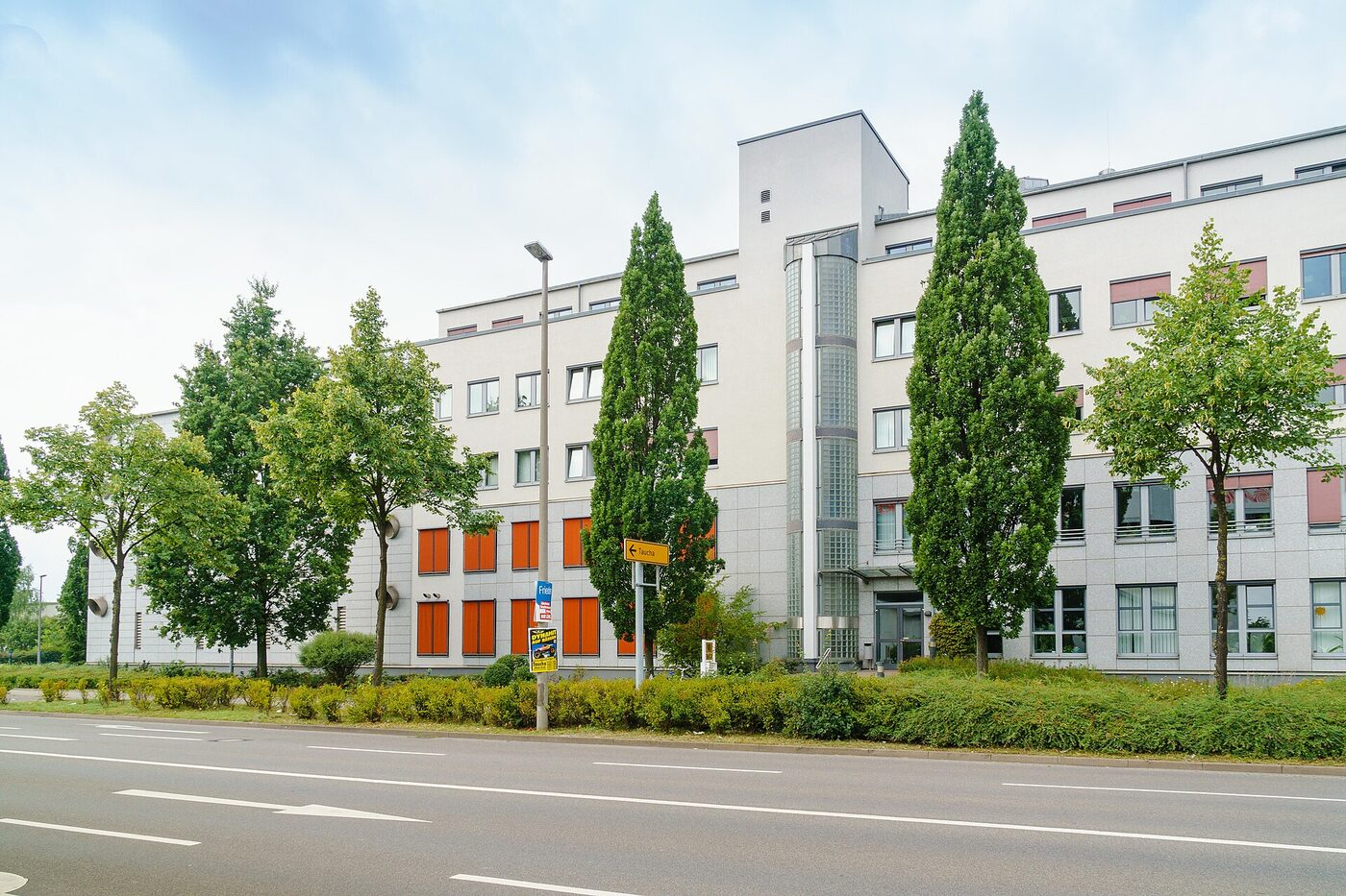 A long four-storey office building in pale stone and render, with a glazed stair tower and orange window panels, seen across a road behind a row of trees on a summer day.
