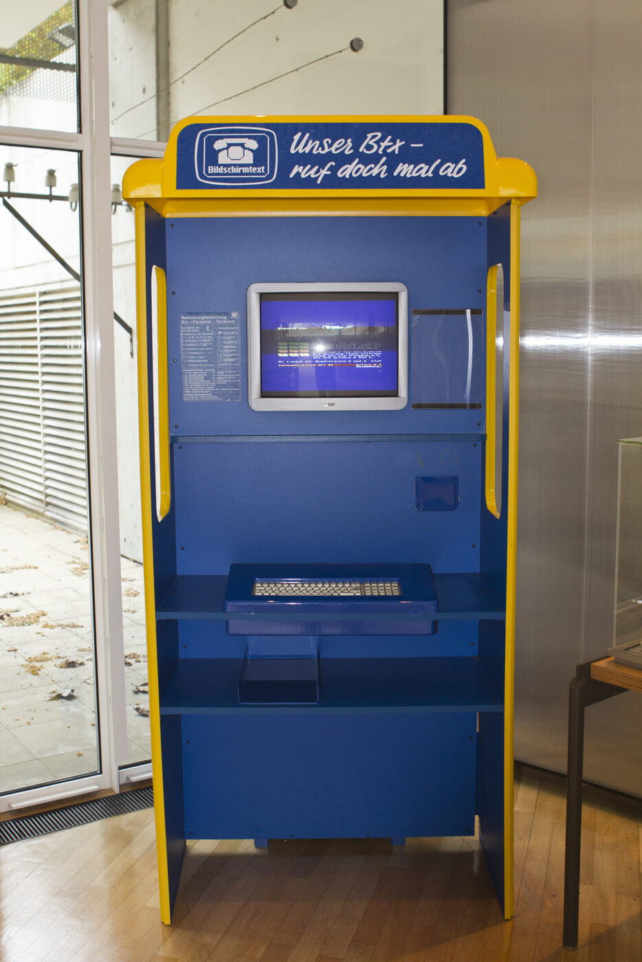 A free-standing public terminal in Bundespost blue and yellow, its header panel lettered Bildschirmtext and the slogan Unser Btx: a colour screen showing a videotex page is set into the upper panel, with a keyboard on a shelf below, standing on a wooden museum floor.
